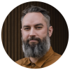Bearded man in a brown shirt against a dark striped backdrop. Ross Featherstone
